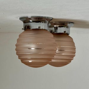 May include: Two flush mount ceiling lights. Each light has a chrome base and a pink, ribbed glass shade. The shades are a soft, muted pink color. The lights are mounted on a white ceiling.