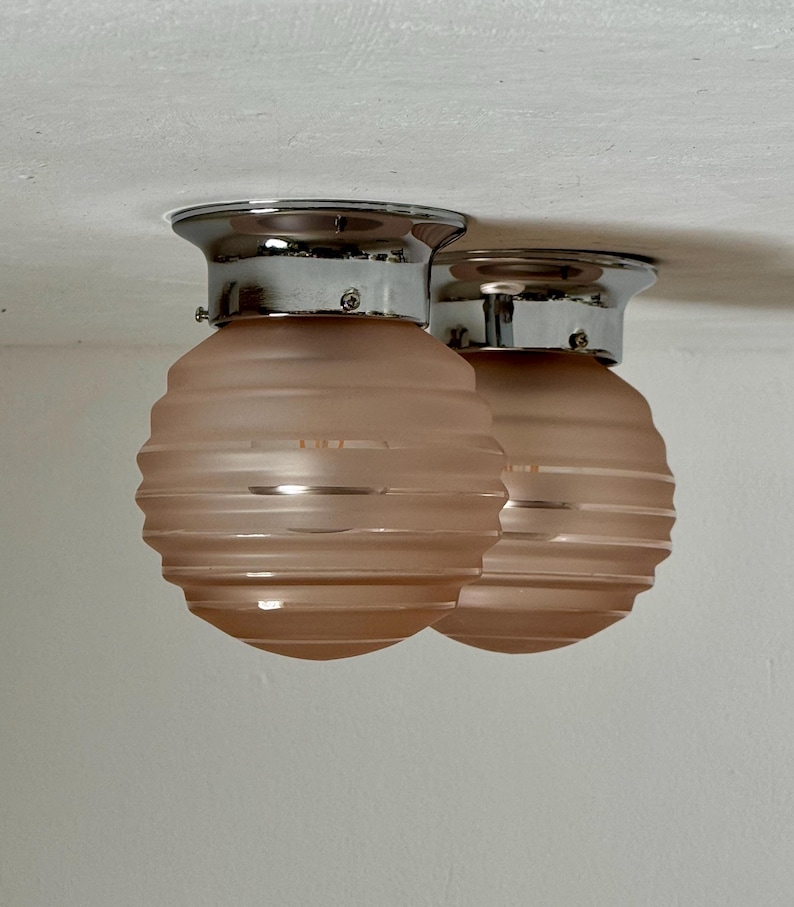 May include: Two flush mount ceiling lights. Each light has a chrome base and a pink, ribbed glass shade. The shades are a soft, muted pink color. The lights are mounted on a white ceiling.