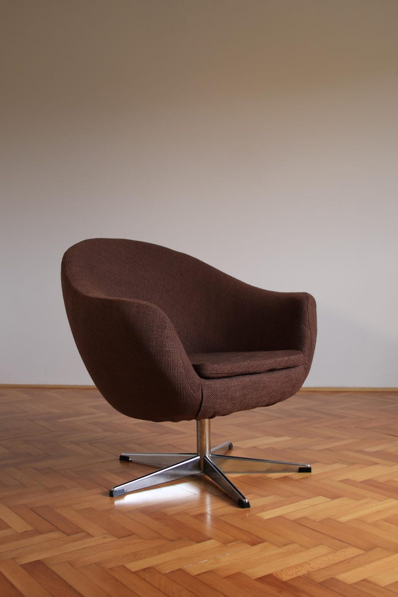May include: A brown, mid-century modern swivel chair with a curved back and seat. The chair has a textured fabric and sits on a chrome, star-shaped base. The chair is positioned on a wooden floor.