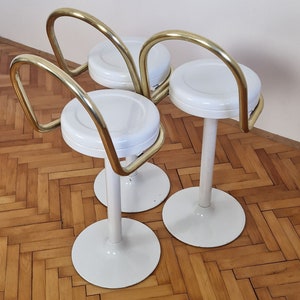 May include: Three white bar stools with gold metal frames. The stools have round seats and a curved back.