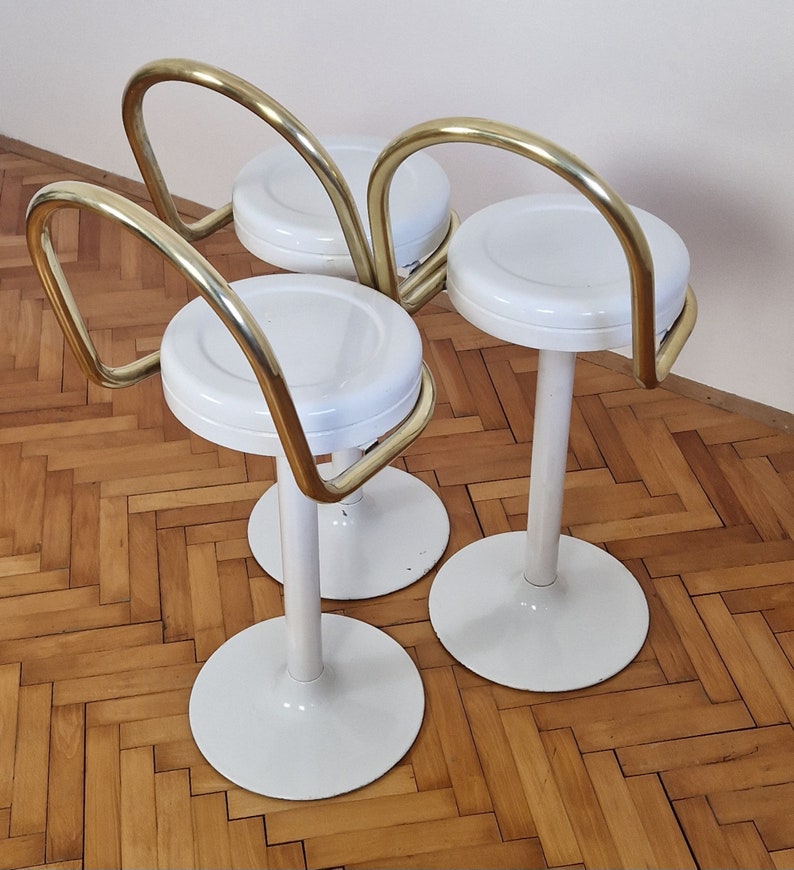 May include: Three white bar stools with gold metal frames. The stools have round seats and a curved back.