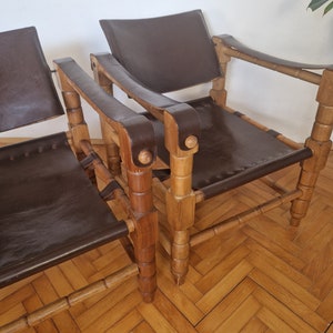 May include: Two brown leather chairs with wooden frames. The chairs have a simple design with a curved back and armrests. The chairs are made of natural wood and have a rustic look.