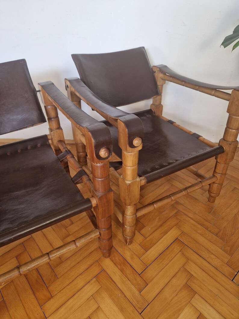 May include: Two brown leather chairs with wooden frames. The chairs have a simple design with a curved back and armrests. The chairs are made of natural wood and have a rustic look.