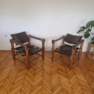 May include: Two brown leather chairs with wooden frames. The chairs have a simple design with a rectangular seat and back, and four legs. The chairs are sitting on a wooden floor.
