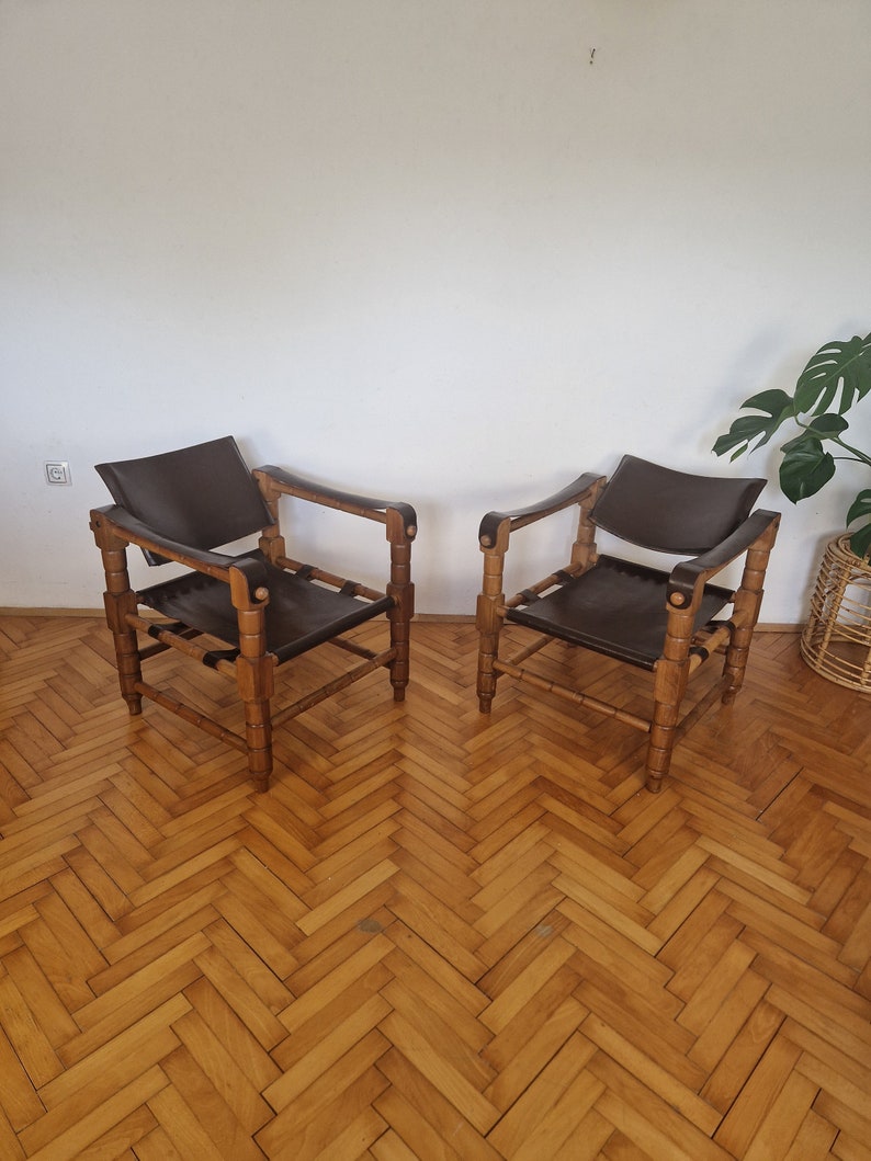 May include: Two brown leather chairs with wooden frames. The chairs have a simple design with a rectangular seat and back, and four legs. The chairs are sitting on a wooden floor.