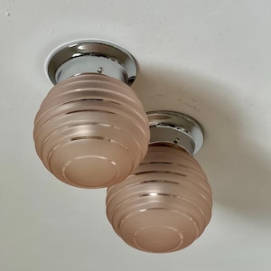 May include: Two vintage ceiling light fixtures. Each fixture has a chrome base and a pink, ribbed glass shade. The shades are egg-shaped and have a frosted appearance. The lights are mounted on a white ceiling.