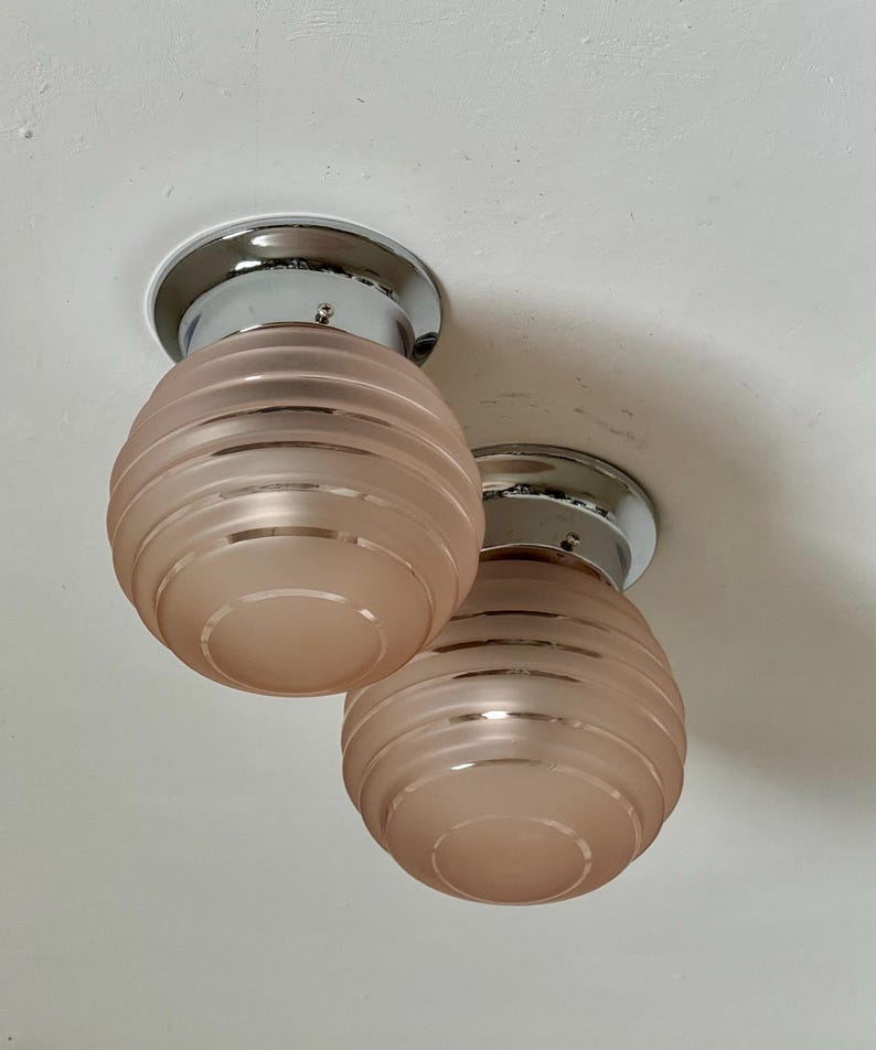 May include: Two vintage ceiling light fixtures. Each fixture has a chrome base and a pink, ribbed glass shade. The shades are egg-shaped and have a frosted appearance. The lights are mounted on a white ceiling.