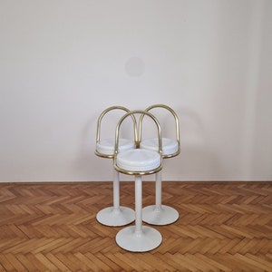May include: Three white bar stools with gold metal frames and round seats. The stools are arranged in a triangular formation on a wooden floor.