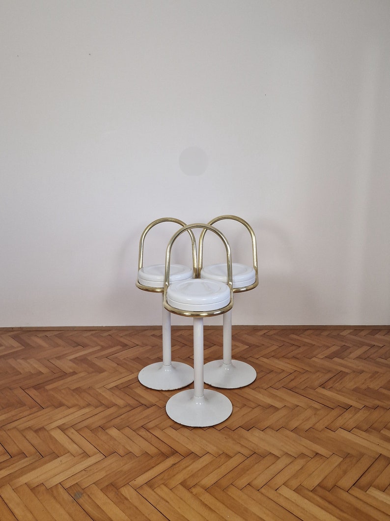 May include: Three white bar stools with gold metal frames and round seats. The stools are arranged in a triangular formation on a wooden floor.