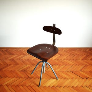Puede incluir: Una silla giratoria industrial vintage con un asiento de madera marr&oacute;n oscuro y un respaldo de metal. La silla tiene una base de metal azul con cinco patas.