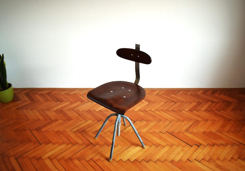 Puede incluir: Una silla giratoria industrial vintage con un asiento de madera marr&oacute;n oscuro y un respaldo de metal. La silla tiene una base de metal azul con cinco patas.
