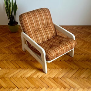May include: A white armchair with a brown and beige striped fabric cushion. The chair has a simple design with rounded edges and wooden arms.