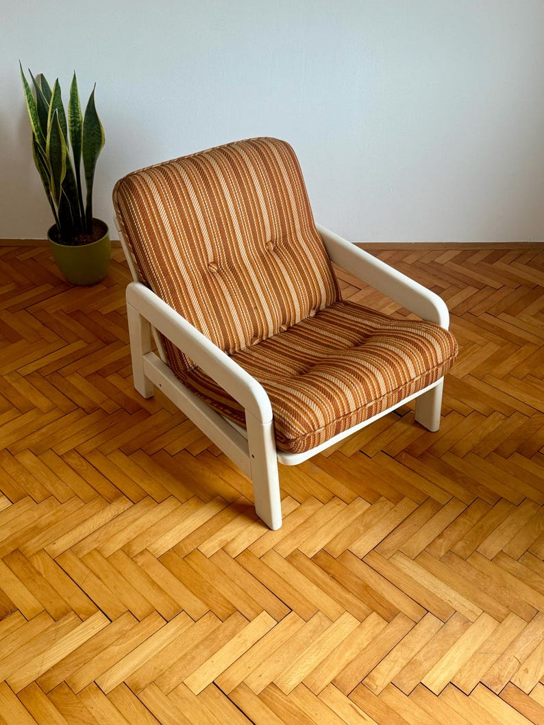 May include: A white armchair with a brown and beige striped fabric cushion. The chair has a simple design with rounded edges and wooden arms.