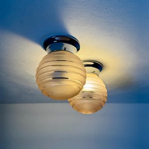 May include: Two ceiling-mounted light fixtures with chrome bases and frosted glass shades. The shades are spherical with horizontal ribbed detailing. The lights are illuminated, casting a warm glow against the white ceiling.