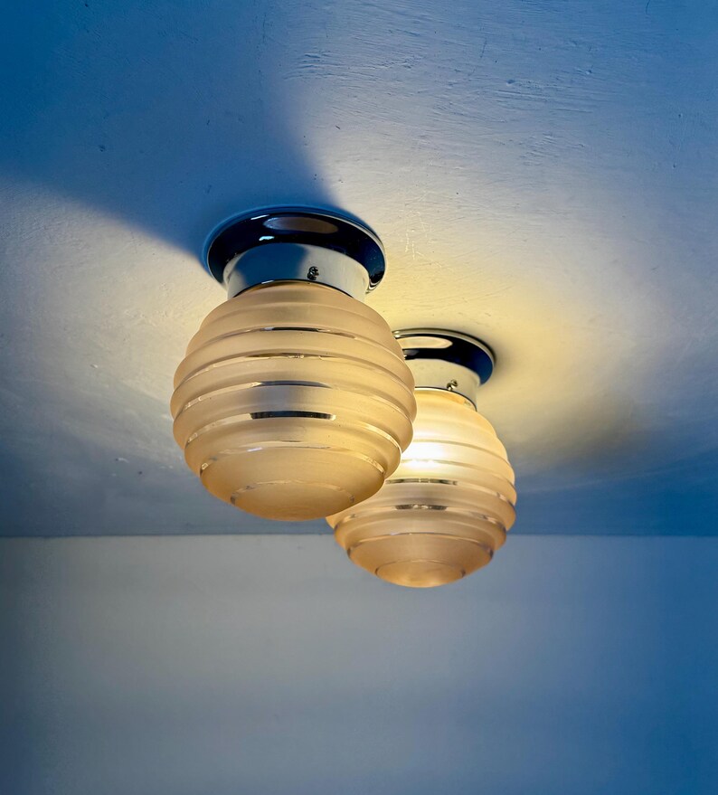 May include: Two ceiling-mounted light fixtures with chrome bases and frosted glass shades. The shades are spherical with horizontal ribbed detailing. The lights are illuminated, casting a warm glow against the white ceiling.