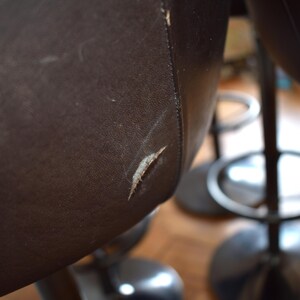 Op de afbeelding: Close-up van een bruine leren barstoel met een kleine scheur in het leer. De stoel heeft een zwarte metalen basis en voetensteun.