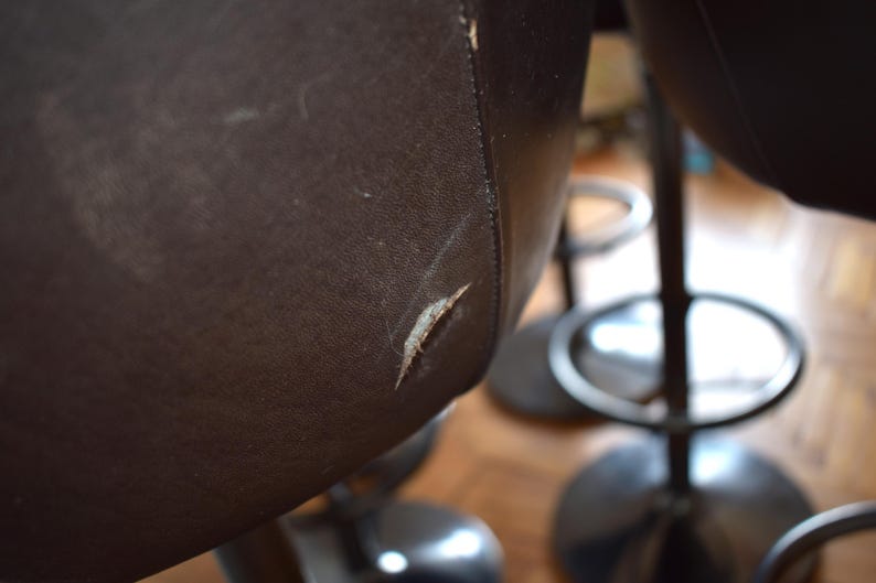 Op de afbeelding: Close-up van een bruine leren barstoel met een kleine scheur in het leer. De stoel heeft een zwarte metalen basis en voetensteun.
