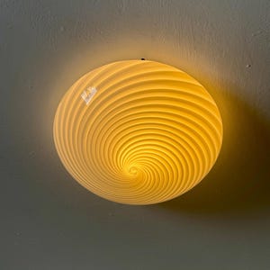 Puede incluir: Un plafón redondo de color crema con un diseño en espiral. La luz está encendida, emitiendo un cálido resplandor amarillo. La lámpara está montada en un techo gris.