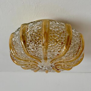 Pode incluir: Uma luminária de teto vintage de vidro transparente com um padrão texturizado semelhante a bolhas. A luminária tem acabamento amarelo dourado e formato arredondado e orgânico. É projetada para ser montada no teto.