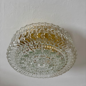 May include: A clear glass ceiling light fixture with a textured, bubble-like pattern. The light has a gold-colored interior and is mounted on a white wall. The fixture is round and has a decorative design.