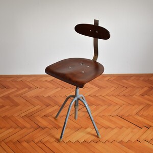 Puede incluir: Una silla giratoria industrial vintage con asiento y respaldo de madera marr&oacute;n, y una base de metal gris con tres patas.