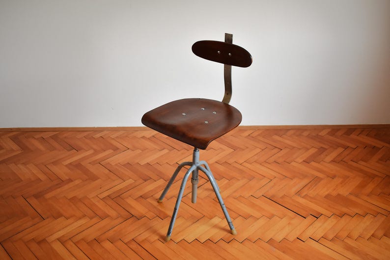 Puede incluir: Una silla giratoria industrial vintage con asiento y respaldo de madera marr&oacute;n, y una base de metal gris con tres patas.