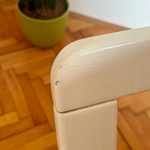 May include: Close-up of a white painted wooden chair with a distressed finish. The chair has a simple design with a square back and legs. The paint is chipped and cracked, giving the chair a vintage look.