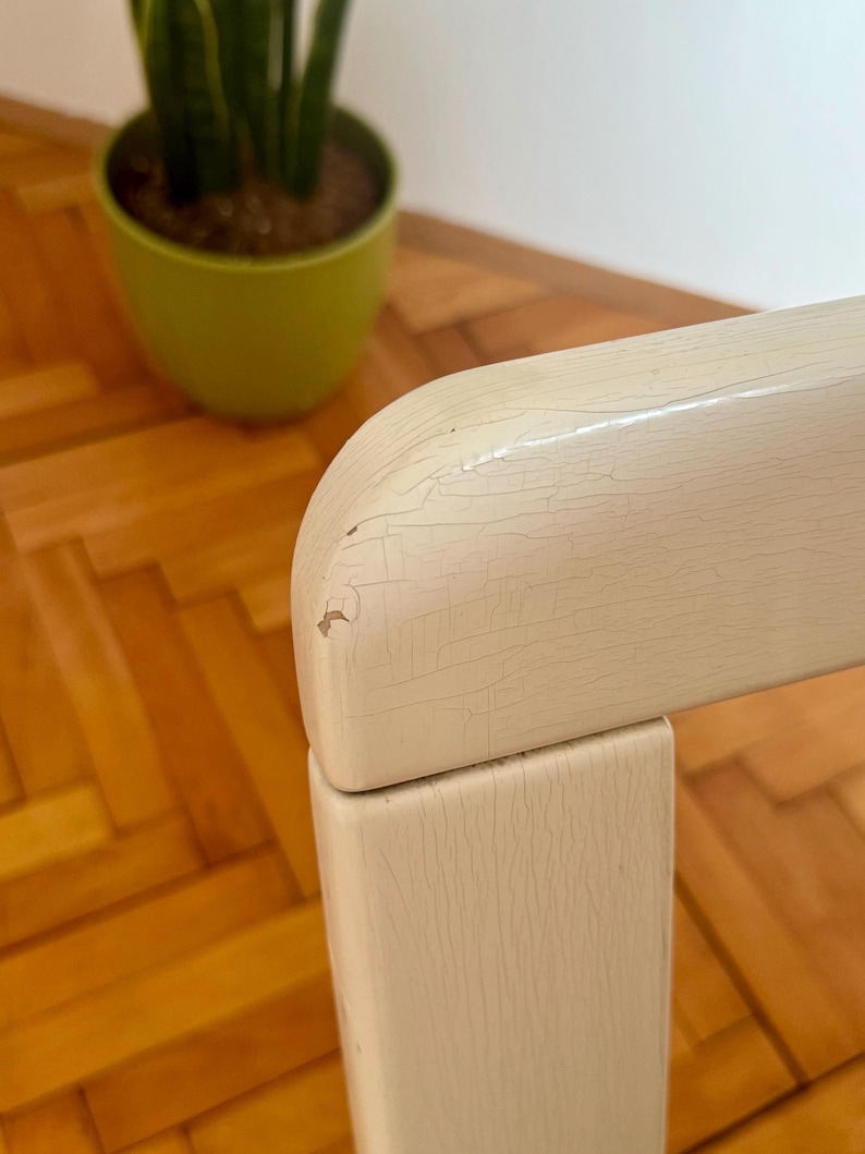 May include: Close-up of a white painted wooden chair with a distressed finish. The chair has a simple design with a square back and legs. The paint is chipped and cracked, giving the chair a vintage look.