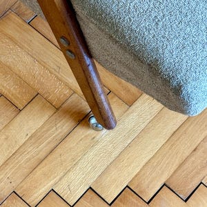 May include: Close-up of a wooden chair leg with a metal caster wheel. The chair is upholstered in a light gray fabric and is sitting on a wooden floor.