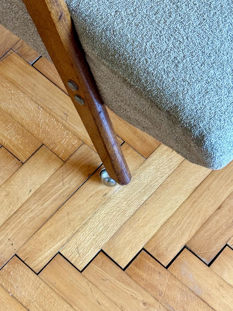 May include: Close-up of a wooden chair leg with a metal caster wheel. The chair is upholstered in a light gray fabric and is sitting on a wooden floor.