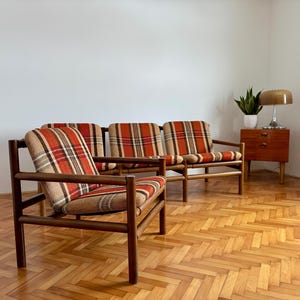 May include: Three mid-century modern armchairs with plaid upholstery in shades of orange, red, and beige. The wooden frames have a dark finish. A small wooden side table with a lamp and a plant are in the background.