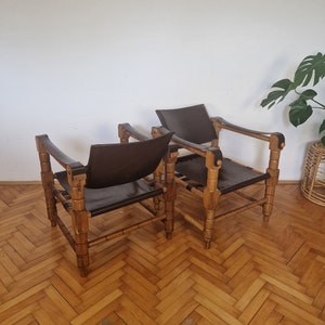 May include: Two brown leather chairs with wooden frames. The chairs have a simple design with a wide, flat seat and a low back. The chairs are made of natural materials and have a rustic look.