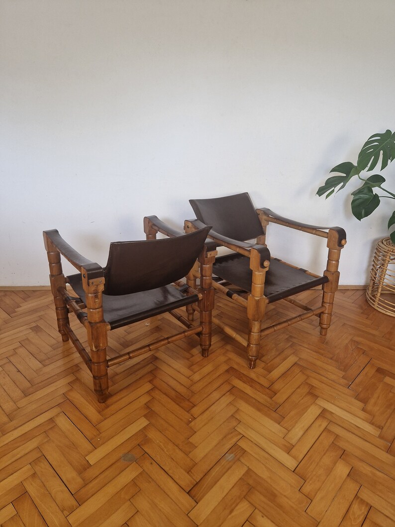 May include: Two brown leather chairs with wooden frames. The chairs have a simple design with a wide, flat seat and a low back. The chairs are made of natural materials and have a rustic look.