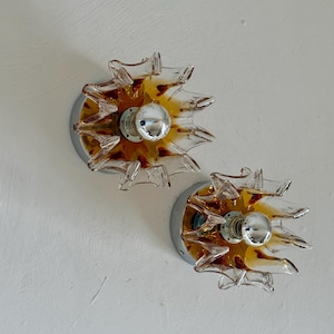 Può includere: Due applique da parete vintage con un design a forma di stella. Ogni applique presenta una base cromata e un paralume in vetro trasparente con accenti ambra e marroni. Le lampadine sono visibili al centro.