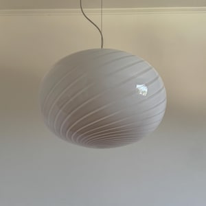 Puede incluir: Una lámpara colgante esférica de vidrio blanco con un sutil patrón de costillas en espiral que envuelve el globo. La lámpara está suspendida del techo por un cable fino de color claro, creando una estética limpia y sencilla.