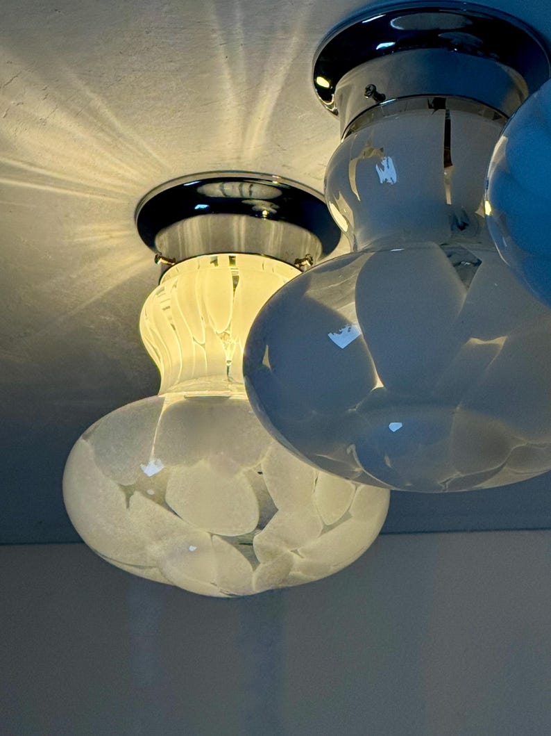 May include: Close-up of a ceiling light fixture with three glass globes. The globes are white with a speckled pattern and are attached to a chrome base. The light is on, illuminating the room.