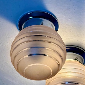 May include: Two Art Deco-style ceiling lights. Each light features a frosted glass globe with horizontal silver bands, mounted on a chrome base. The lights are suspended from a white ceiling, creating a vintage aesthetic.