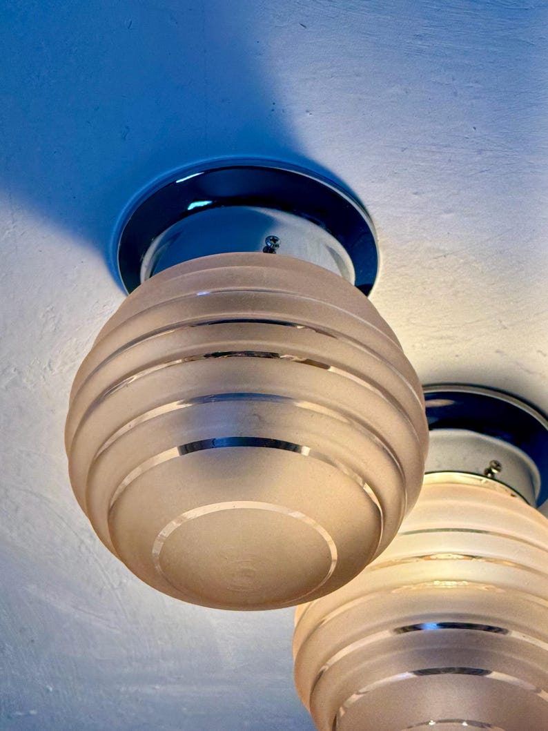 May include: Two Art Deco-style ceiling lights. Each light features a frosted glass globe with horizontal silver bands, mounted on a chrome base. The lights are suspended from a white ceiling, creating a vintage aesthetic.