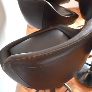 Op de afbeelding: Een set van bruine leren barstoelen met zwarte metalen bases. De krukken hebben een gebogen rugleuning en een comfortabele zitting.