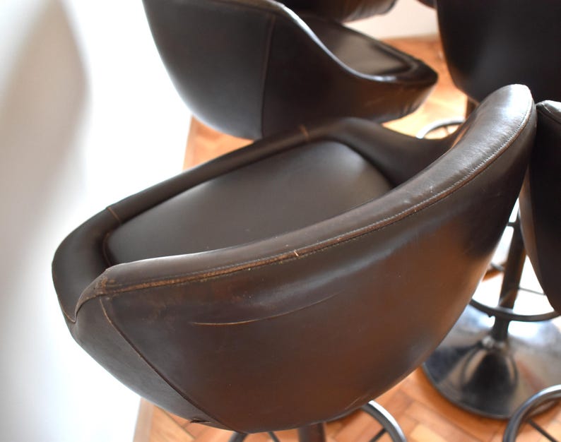Op de afbeelding: Een set van bruine leren barstoelen met zwarte metalen bases. De krukken hebben een gebogen rugleuning en een comfortabele zitting.