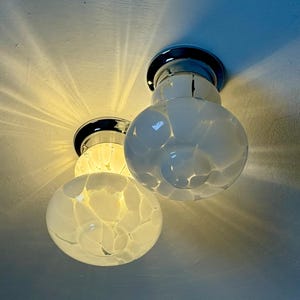 May include: Two modern ceiling lights with a chrome base and white glass globes. The globes have a textured, bubble-like pattern. The lights are illuminated, casting a warm glow and shadows on the ceiling.