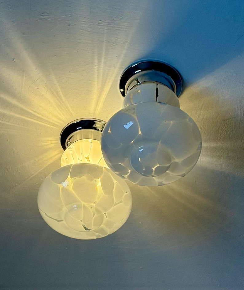May include: Two modern ceiling lights with a chrome base and white glass globes. The globes have a textured, bubble-like pattern. The lights are illuminated, casting a warm glow and shadows on the ceiling.