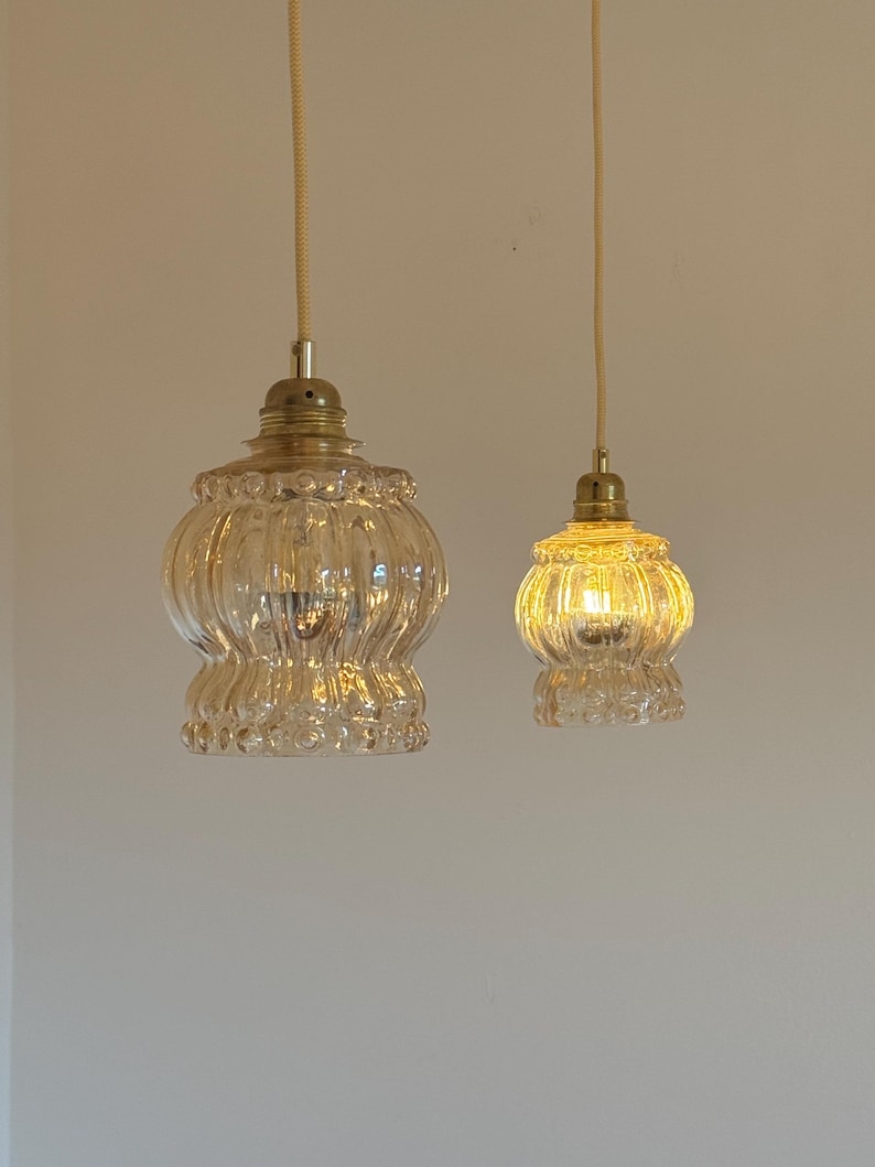 May include: Two clear glass pendant lights with a ribbed texture hang from a white ceiling. The larger light has a wider, more rounded shape, while the smaller light is more spherical. Both lights have a brass finish.