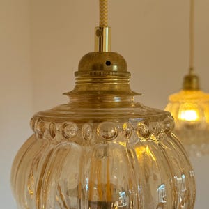 May include: A close-up of a gold-toned pendant light with a clear, amber-colored glass shade. The shade has a textured, ribbed design and a beaded rim.