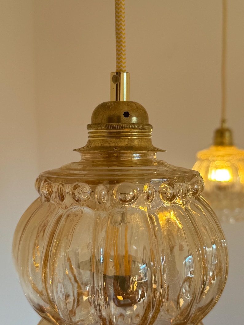 May include: A close-up of a gold-toned pendant light with a clear, amber-colored glass shade. The shade has a textured, ribbed design and a beaded rim.