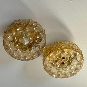 May include: Two vintage, amber-colored glass light fixtures. Each fixture is a rounded dome shape with a floral pattern. The glass has a textured surface, and the fixtures are likely for ceiling or wall mounting.