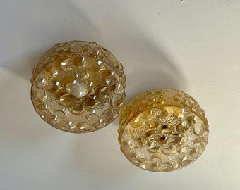 May include: Two vintage, amber-colored glass light fixtures. Each fixture is a rounded dome shape with a floral pattern. The glass has a textured surface, and the fixtures are likely for ceiling or wall mounting.