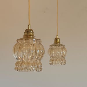 May include: Two clear glass pendant lights with a ribbed texture and gold metal accents hang from white cords against a white wall.