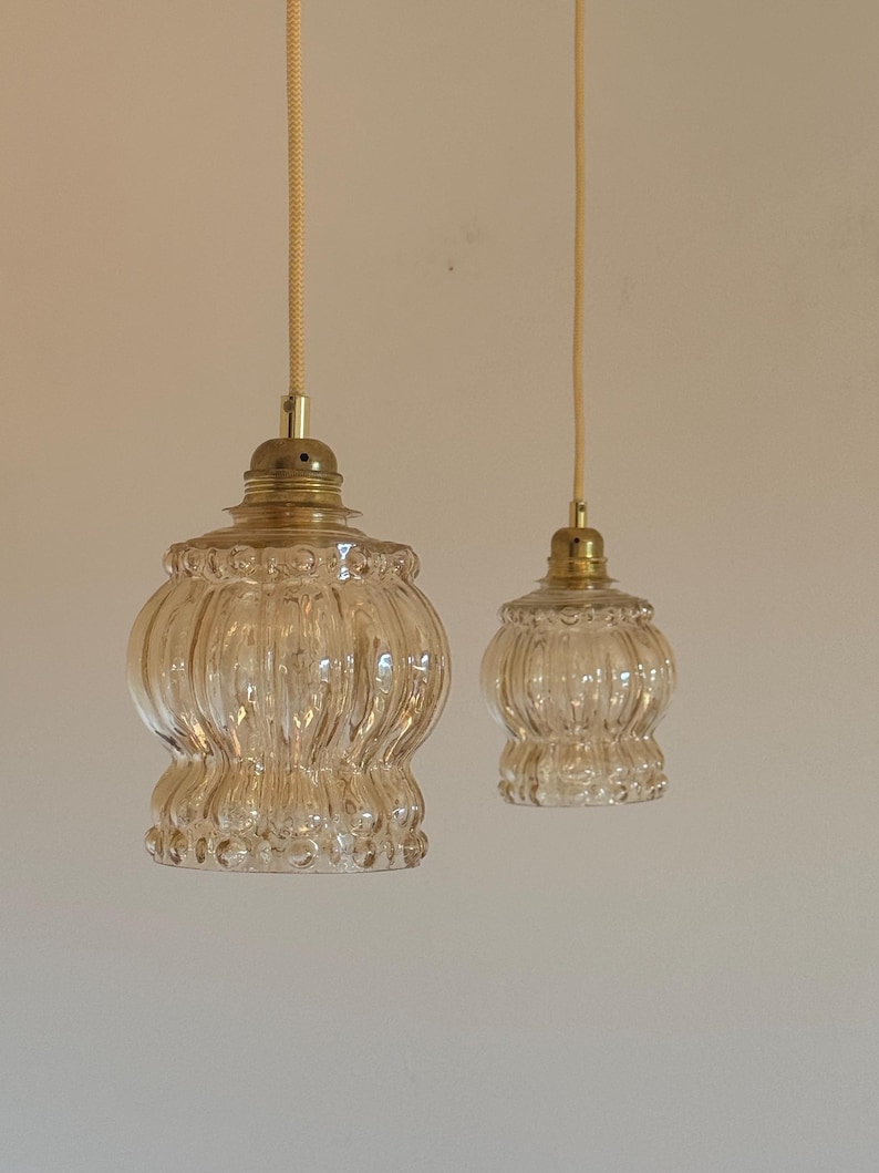 May include: Two clear glass pendant lights with a ribbed texture and gold metal accents hang from white cords against a white wall.