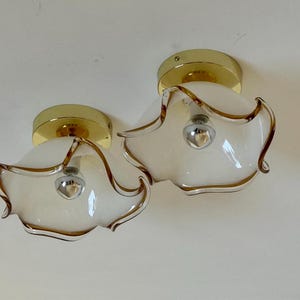May include: Two vintage ceiling light fixtures. Each fixture has a gold-colored base and a white glass shade with a brown trim. The shades have a unique, wavy shape, and the light bulbs are visible.